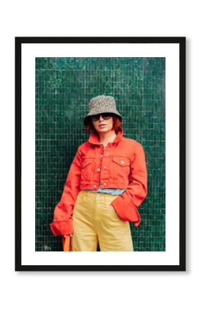 Hipster fashion young woman in bright clothes, sun glasses and bucket hat posing on the green tile wall background. Urban city street fashion. Vertical card. Selective focus. Copy space.