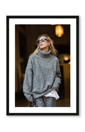 blonde woman in stylish sunglasses and grey sweater standing with black handbag in New York