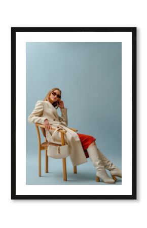 Fashionable confident woman wearing elegant white woolen coat, sunglasses, high leather heeled boots, posing on blue background. Full-length studio fashion portrait. Copy, empty space for text