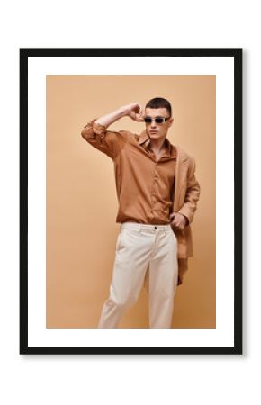Classy man in beige jacket on shoulder, shirt, pants and sunglasses posing on beige background