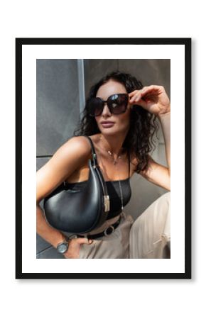Glamorous beautiful young urban fashion woman with curly hairstyle in fashionable clothes with top and pants with stylish handbag puts on sunglasses and sits near building in the city
