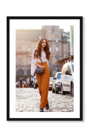 Outdoor full body fashion portrait of young beautiful woman wearing stylish yellow high-waisted wide-leg pants, white blouse, holding green leather bag. Model walking in street of european city