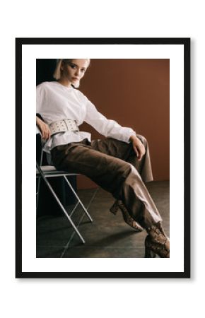 stylish blonde woman in white blouse and boots with snakeskin print sitting on chair near curtain on brown