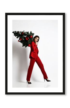 Beautiful fashion brunette girl in a stylish red office jacket suit walks and carries a Christmas tree decorated with balls