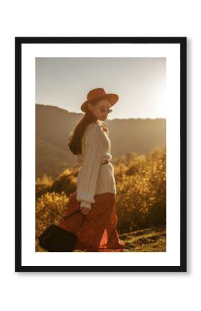 Outdoor autumn fashion portrait of beautiful confident woman wearing stylish hat, sunglasses, dress, knitted sweater, belt, holding leather handbag, posing, walking at sunset in mountain landscape