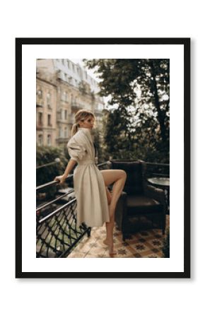 Full height portrait of a stylish blonde woman with long legs in elegant leather trench coat and smoky eyes makeup in the outdoor on the balcony. Spring - autumn fashion concept. Soft selective focus.