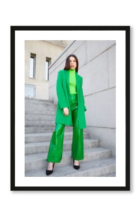 High fashion photo of a young brunette woman in green coat, poloneck and leather pants.