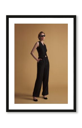 Elegant woman in a sleeveless black vest and wide-leg trousers posing with sunglasses against a beige background. Stylish, powerful, modern fashion concept.