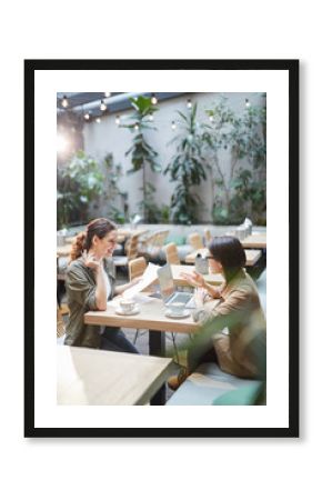High angle portrait of two modern young women sitting at table in designer cafe and smiling cheerfully while discussing business, copy space