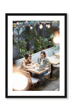 High angle portrait of two young women working together sitting at table in outdoor cafe terrace decorated with plats, copy space