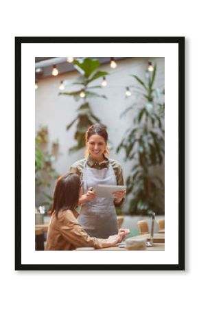 Portrait of smiling young waitress holding digital tablet while taking orders at outdoor cafe terrace lit by warm sunlight
