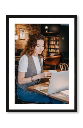 A young woman with curly red hair enjoys a coffee while working on her laptop in a warm cafe space. Freelance life, mobile business, casual professionalism, on-the-go workflow...