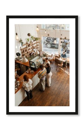 Adults inside a modern cafe with wooden floors and large windows, some standing in line to order from the barista at the counter while one man sits by the window enjoying a drink.