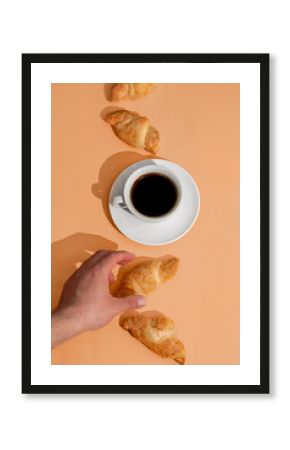 Hand holding fresh croissant on orange background with coffee cup and fresh pastry on plate. Creative layout for cafe advertising or French breakfast concept. Top view.