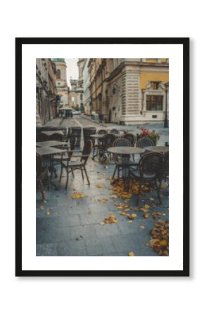 Empty outdoor café with wooden chairs and tables on a stone street, covered with fallen autumn leaves in a historic European city center