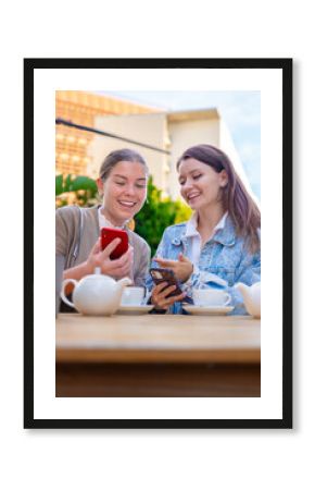 Two happy friends women sitting at an outdoor cafe, using smartphones and reacting with curiosity and surprise. Surrounded by teapots and cups, they share a modern and relaxed social moment together. 