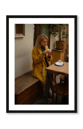 Young woman in an orange coat sitting at a cozy café table with a cup of coffee, gazing dreamily into the distance. Warm autumn atmosphere, urban lifestyle, mindful moment.