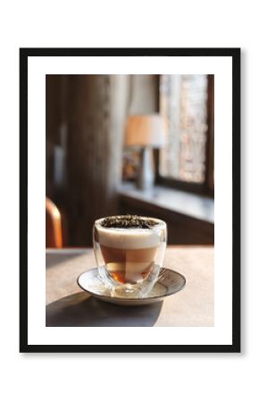 Dry tea leaves scattered artistically over the foamy top of a transparent latte glass. Eye-level view, light streaming from side window, clean table with soft ceramic patterns, cozy indoor café feel.