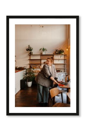 A white woman wearing a blazer uses a laptop while standing inside a modern cafe during renovation or setup, surrounded by wooden furniture, plants, and shelves.