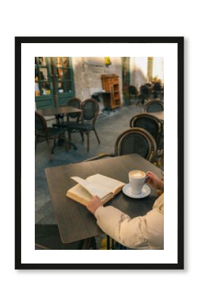A person enjoys a latte and a book at an outdoor café table on a quiet street. Cozy autumn or winter vibe with warm light and European-style architecture in the background