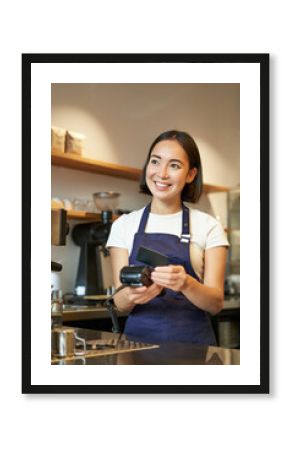 Portrait of smiling asian barista, coffee shop employee using POS terminal and credit card, helps client pay contactless in cafe