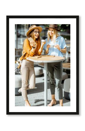 Two female best friends spending time together on the cafe terrace, feeling happy standing with coffee and phone during a summer day