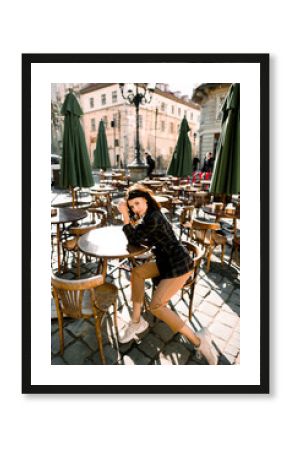 fashion outdoor photo of beautiful woman with dark hair in elegant modern clothes posing while sitting on wooden chair in city cafe outdoors, old city street and buildings on the background