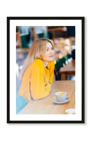 Portrait of blond woman in yellow pullover sits with cup of coffee or tea at the beautiful loft interior of the cafe on background.