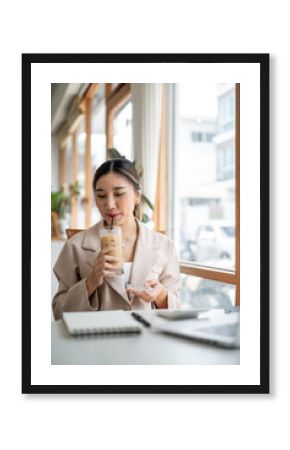 Asian woman holding glass coster drinking ice coffee with straw as looking at notebook on cafe table