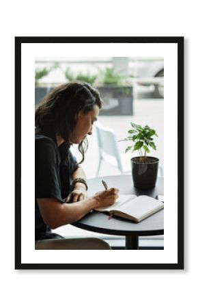Vertical medium side view portrait shot of beautiful young Caucasian woman sitting at cafe table writing something in her notebook