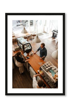 An overhead view of a mid-30s White man ordering coffee while standing in a small coffee shop, as a young, White woman in her 20s stands behind the counter, with her male colleague making the order