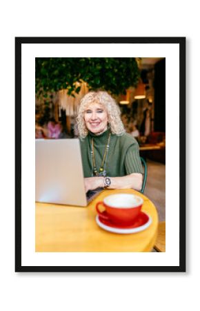 Happy senior woman in green sweater smiling at camera while using laptop in bright cafe. Concept: senior empowerment, age inclusivity, digital skills.