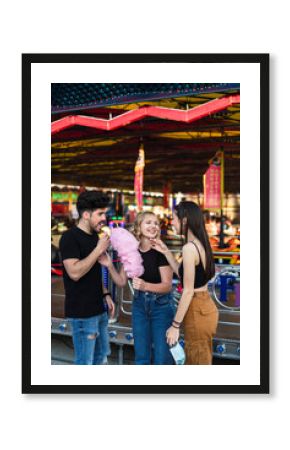 Dos chicas y un chico amigos sonriendo y compartiendo un algodón de azúcar rosa en zona de atracciones de una feria