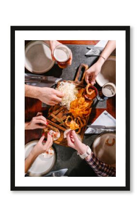 A company of friends of different sexes sits in a pub with beer snacks on a wooden board. Potato and rice chips, croutons, onion rings, smoked cheese, tomato sauce, tartar sauce and beer.