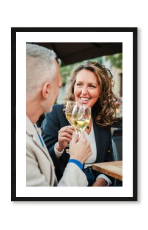 Vertical. Mature couple toasting white wine glasses on a bar terrace. Middle aged married people drinking alcohol on a restaurant. Mid adult friends clinking with alcohol beverages on a date at pub