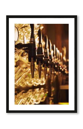 Vintage vibe of bar. Close-up of multiple shiny beer taps with handles, set against blurred background with rows of empty glasses hanging. Concept of beer, brewery, pub, Oktoberfest, traditions