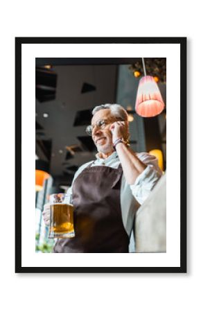 senior man in apron talking on smartphone and holding glass of beer in pub