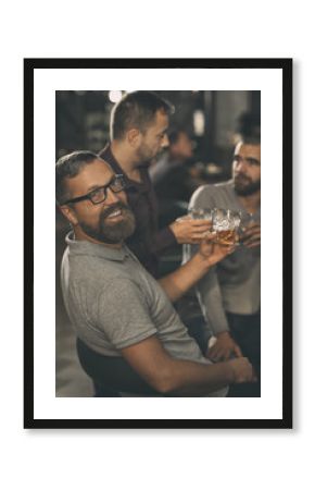 Side view of positive man wearing in spectacles and grey t shirt looking at camera and smiling. Bearded man holding glass of scotch. His friends standing behind and communicating.