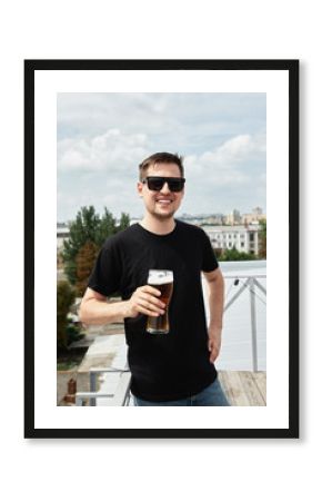 Happy man in black shirt and eyewear drinking beer at bar or pub on roof, copy space