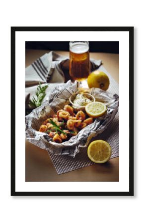 Close-up of crispy calamari rings served with dipping sauces, lemon slice, and herbs on newspaper-style paper, with a glass of beer in background