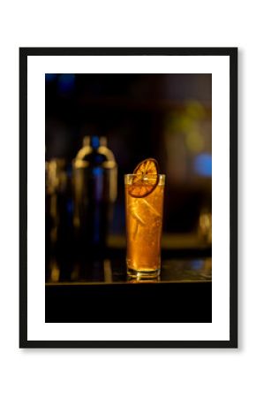 Amber cocktail in highball glass, garnished with dried orange slice, resting on dark bar counter near cocktail shakers, illuminated by soft lighting