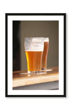 Two glasses of beer one of full and the other is just over half full. They both have coasters and are just sitting on a bar table.