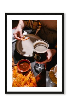 A girl drinks dark beer in a pub with beer snacks on a wooden board. Potato and rice chips, croutons, onion rings, smoked cheese, tomato sauce, tartar sauce.
