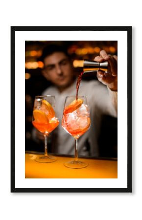 glass with ice cubes and orange slices on the bar and bartender pours drink from jigger into glass