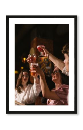 Happy people cheers glasses with cocktails celebrating event in bar. Joyful friends raise up drinks at party in night club