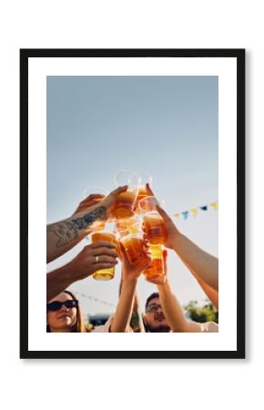 Group of people, friends gathering on picnic, raising glasses with beer, clinking under blue sunny clear sky. Traditional festival. Concept of summer, leisure, friendship, meeting, fun, relaxation