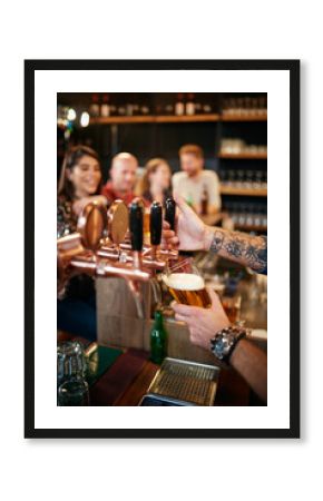 Cropped picture of tattooed barman pouring beer in glass. In background is group of friends sitting at counter. Pub interior.