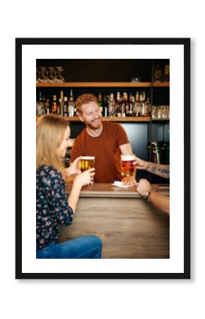 Cheerful friends leaning on bar counter, drinking beer and chatting with bartender. Night out.