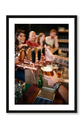 Cropped picture of tattooed barman pouring beer in glass. In background is group of friends sitting at counter. Pub interior.