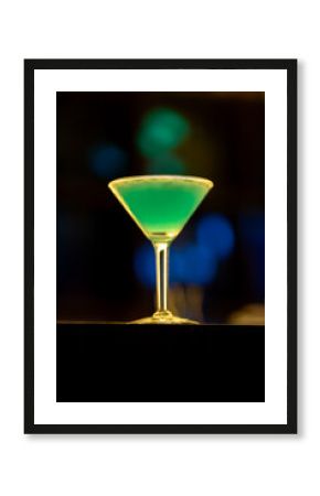 Green cocktail garnished with lime, resting on wooden bar surface, blurred lights highlighting elegant mixology scene